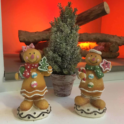 Figuras decorativas de biscoitos de Natal com enfeites coloridos ao redor de uma mini árvore de Natal em vaso, com lareira acesa ao fundo
