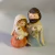 Miniatura de Sagrada Família colorida em tons pastéis, feita em resina, mostrando Maria segurando o menino Jesus e José com uma lanterna