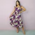 Mulher vestindo vestido midi floral uva com estampa de flores em tons de roxo, lilás e branco, posando com uma mão na cintura e outra segurando a barra do vestido