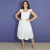Mulher sorridente usando vestido midi off white com detalhes de babados nos ombros, posando em fundo azul