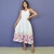 Mulher usando vestido branco mídi com estampa floral colorida na barra, posando com uma mão na cintura e sorrindo