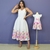 Vestido branco midi com estampa floral colorida na barra usado por mulher ao lado de vestido infantil branco com estampa floral e laço rosa exposto em manequim