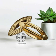 Fivela de metal dourada com design rajado, medindo 5,5cm x 2,5cm, posicionada ao lado de uma planta em vaso.
