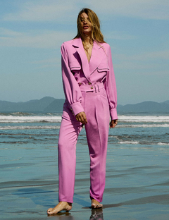 Modelo usando um blazer rosa com botões dourados em uma praia, com o mar ao fundo.