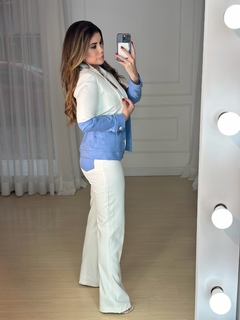 Modelo usando um blazer branco com detalhes em denim azul, posando de lado em um ambiente com iluminação suave.