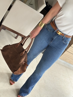 Imagem de uma mulher usando calça flare jeans de cintura média, segurando uma bolsa marrom e exibindo um bracelete dourado.