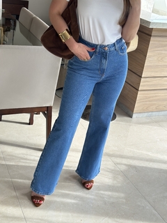 Mulher usando calça jeans wide leg com elastano, na cor azul, combinada com uma camiseta branca e sandálias. Ela está em um ambiente interno elegante.