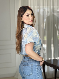Modelo usando uma camisa bordada de manga curta em tom azul, combinada com jeans, em um ambiente iluminado.