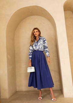 Modelo usando uma saia midi azul com pregas e blusa estampada em fundo claro, segurando uma bolsa branca.