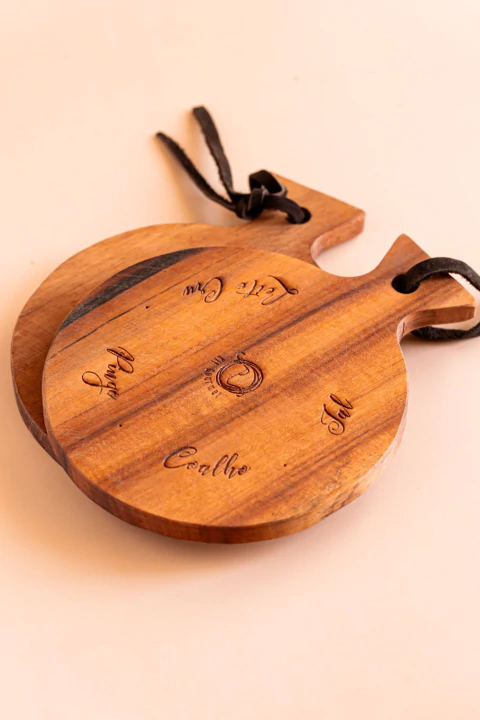 Tábua para queijo artesanal da Provençal Gourmet. Redonda de madeira, com alça e cordão preto, contendo gravações de nomes de queijos.