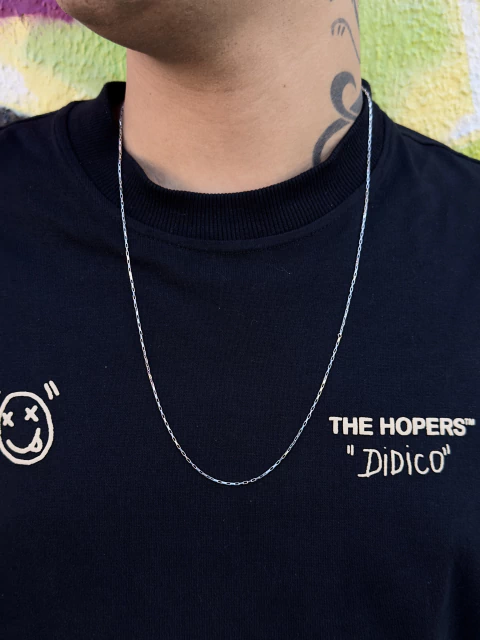 Jovem com cabelo cacheado e colar de corrente tijolinho de prata 925, posando em frente a um mural colorido.