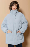 Mulher usando jaqueta feminina Biamar oversized azul clara com gola alta e bolsos frontais, em fundo bege