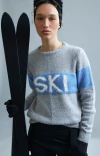 Modelo vestindo blusa ambicione por anselmi cinza com estampa 'SKI' em azul, segurando esquis pretos.