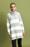 Modelo usando blusa ambicione por anselmi maxi pull cinza com gola alta e listras brancas em fundo verde.