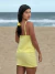 Mulher usando a Saia Maitê Amarela curta com elástico na cintura, ideal como saída de praia, em cenário de praia ao fundo.