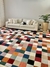 Tapete geométrico com padrão de quadrados coloridos em tons de azul marinho, terracota, off-white, verde, verde claro, bordô, granito, rosé gold, bege, cobre e nude rosado, em uma sala com sofá claro e planta decorativa.