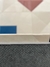 Tapete Optik com fundo off-white e detalhes geométricos coloridos em azul marinho, azul petróleo, bordô, rosé gold e granito, mostrando o verso antiderrapante.