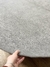 Tapete Bouclé Capri Cinza com bordas orgânicas sendo segurado por uma mão sobre piso de madeira, São José.