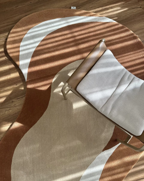 Tapete orgânico da coleção Renzo, confeccionado em nylon Econyl, nas cores trigo, off-white, mel e caramelo, com design exclusivo, posicionado em piso de madeira ao lado de uma poltrona clara.