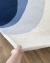 Tapete em nylon com cores azul marinho, azul petróleo, prata e off-white sendo segurado em um ambiente de piso de madeira.