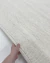 Tapete de fibra natural off-white sendo segurado por uma mão, destacando a textura tramada e o acabamento artesanal do modelo retangular da Coleção Zeland 01, Tapetes São José