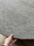 Mão segurando o tapete Bouclé Capri cinza médio com bordas orgânicas, mostrando a textura e acabamento sobre um piso de madeira, São José.