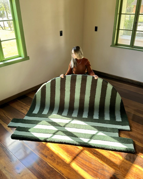 Mulher segurando tapete urbano de nylon com bordas orgânicas, em tons de verde e verde escuro, em uma sala iluminada.