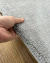 Tapete liso de nylon na cor cinza médio sendo segurado por uma mão, mostrando a textura e espessura sobre um piso de madeira.