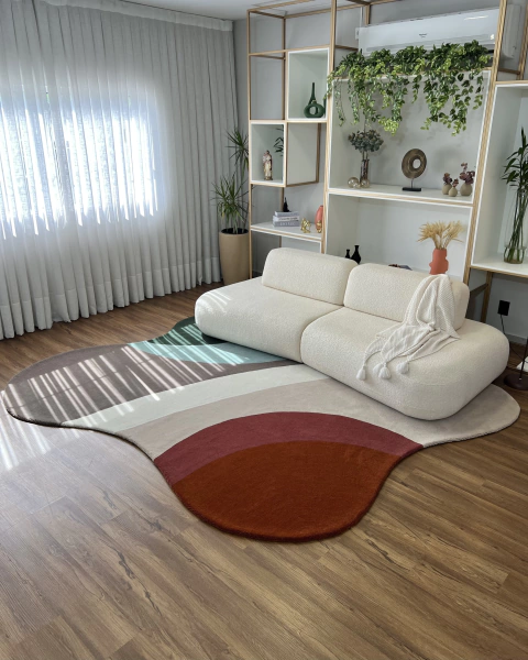 Tapete Kurve® com bordas orgânicas em tons de terracota, cobre, bege claro, off-white, taupe, verde claro e verde Econyl, decorando uma sala de estar moderna com sofá bege e prateleiras ao fundo.