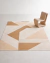 Tapete sala retangular em tons terrosos com padrão geométrico, confeccionado em Bouclé Capri, decorando ambiente minimalista com cadeira moderna bege.