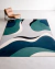 Tapete retangular moderno com padrão ondulado nas cores verde, preto, off-white, cinza e azul, da Coleção Wave, posicionado em uma sala minimalista ao lado de uma poltrona de couro marrom claro.