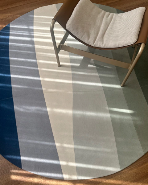 Tapete redondo para sala com design em ondas nas cores azul, verde e cinza, com cadeira de metal e almofada bege sobre ele.
