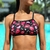 Top cropped de natação com estampa de boquinhas em rosa, vermelho e preto, com raios brancos. Modelo com alças reguláveis em "V" nas costas, usado por uma mulher em frente a uma piscina.