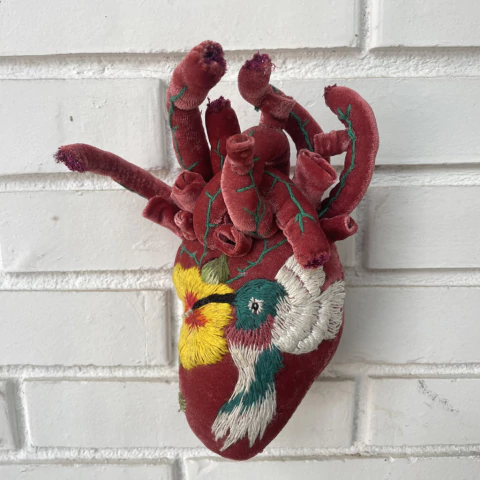 Coração de veludo vermelho com bordados coloridos de flores e pássaro, segurado por uma mão em área externa com grama e plantas ao fundo