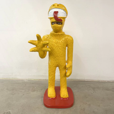 Estátua de uma figura amarela com uma expressão peculiar, segurando a mão em um gesto de saudação.