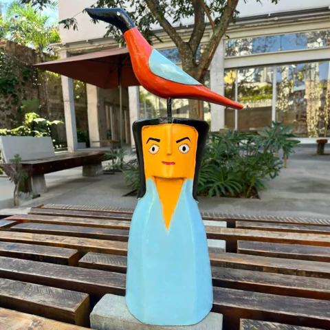 Escultura colorida de uma mulher com um pássaro em cima da cabeça, em um ambiente ao ar livre.