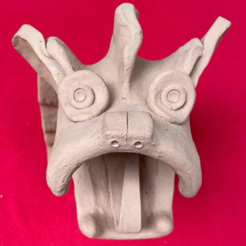 Escultura de carranca em cerâmica natural com olhos redondos e boca aberta, sobre fundo vermelho