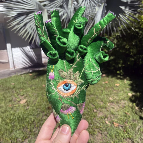 Coração decorativo de veludo verde com bordados de olho azul e flores, em formato anatômico, segurado por uma mão ao ar livre