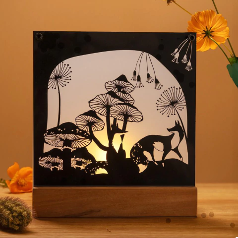 Iluminária decorativa em formato de raposa, com silhuetas de cogumelos e flores, iluminada por trás.