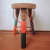 Banco artesanal colorido com um design que representa uma figura humana, com braços levantados e detalhes em várias cores.