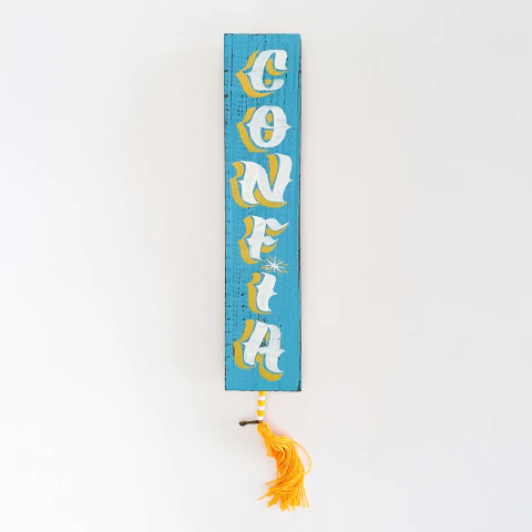 Escultura em madeira pintada de azul com a palavra "CONFIA" em letras brancas e amarelas, com um tassel amarelo pendurado na base