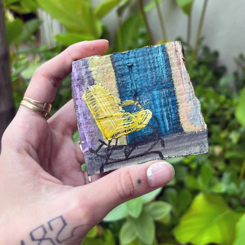 Pintura em madeira de uma cadeira amarela em frente a uma porta azul, segurada por uma mão.