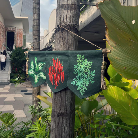 Bandeira decorativa verde com pintura de folhas e pimentas vermelhas pendurada em um tronco de árvore em ambiente externo