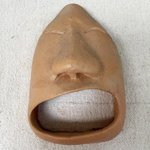 Máscara de cerâmica em formato de rosto humano com olhos fechados e boca aberta, feita de argila.