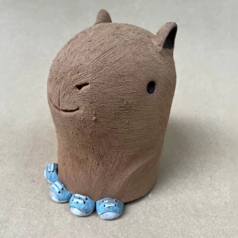 Escultura de cerâmica representando uma capivara com expressão facial simpática, feita por Carlos Lima, com detalhes texturizados e pequenos elementos azuis na base.