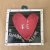 Quadro com coração vermelho e a palavra "Fé" escrito em letras brancas, acompanhado da frase "O amor é assim".