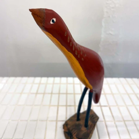 Pássaro decorativo de madeira, pintado em tons de vermelho e amarelo, com pernas azuis, representando o Bento de Sumé.