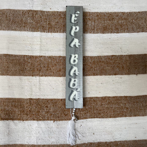 Placa decorativa em madeira com a palavra "PAPABABÁ" em letras brancas tridimensionais, com fita de tassel na parte inferior, sobre fundo listrado em tons de marrom e branco.