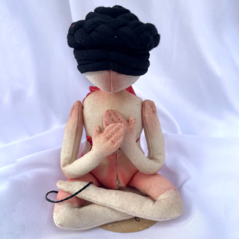 Boneca artesanal sentada de costas com cabelo preto trançado e mãos cruzadas sobre o peito