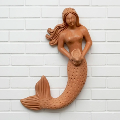 Escultura de uma sereia de cerâmica, com cabelos longos e corpo estilizado, segurando uma concha, fixada em uma parede branca.